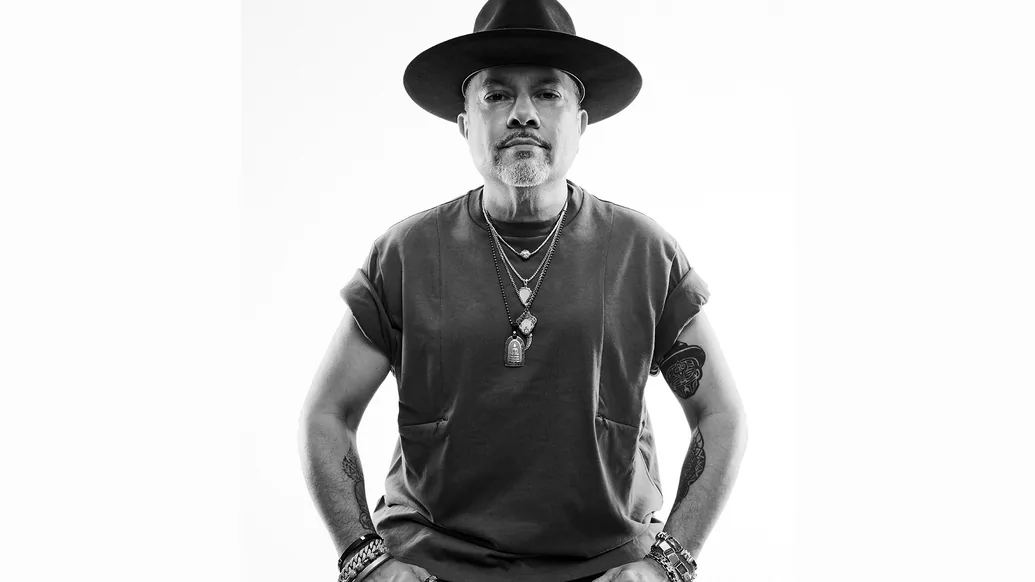 Black and white photo of Louie Vega wearing a black hat and t-shirt with his hands in his pockets
