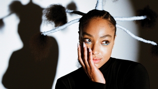 Photo of HoneyLuv under a spotlight wearing a black turtleneck and vertical braids in her hair 