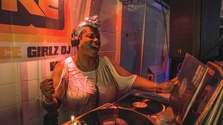 Photo of Marcia Carr DJing on vinyl, holding a record sleeve in one hand and singing passionately