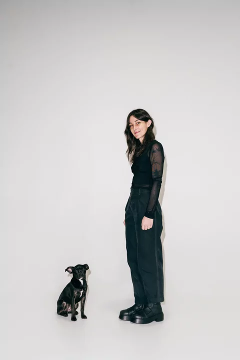 Photo of DJ Voices standing in front of an off-white wall. There is a small, cute black dog beside her