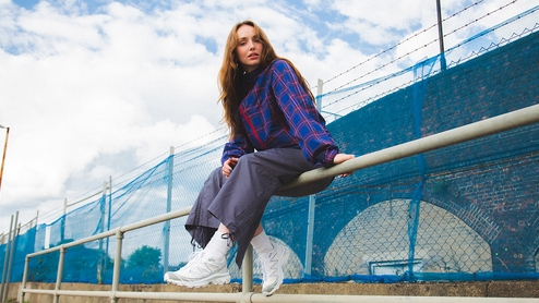 LUXE sitting on a steel bar fence in front of a blue mesh fence near some railway arches. She's wearing a dark blue top with red horizal and vertical stripes, grey blue trousers and white trainers