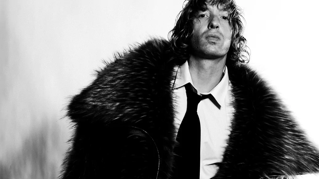 Daniel Avery in black and white looking at the camera in a big fur coat, white shirt and black tie