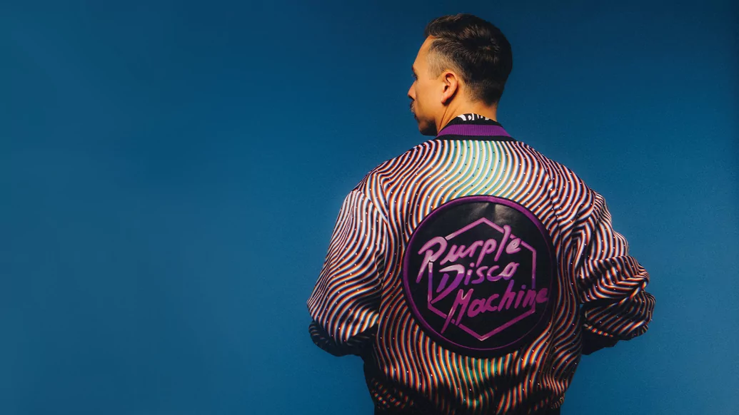 Purple Disco Machine standing with his back to the camera, wearing a jacket with his name emblazoned on it in purple font