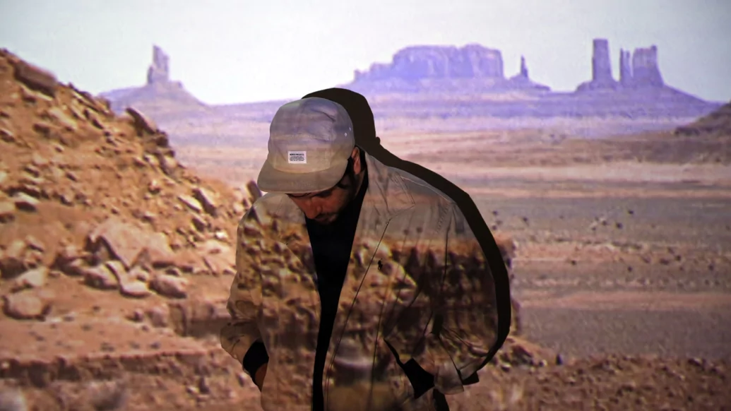 Cover image from from John Talabot's DJ-Kicks mix. He's standing in front of a projection of a desert valley and wearing a beige jacket and hat. He's looking down