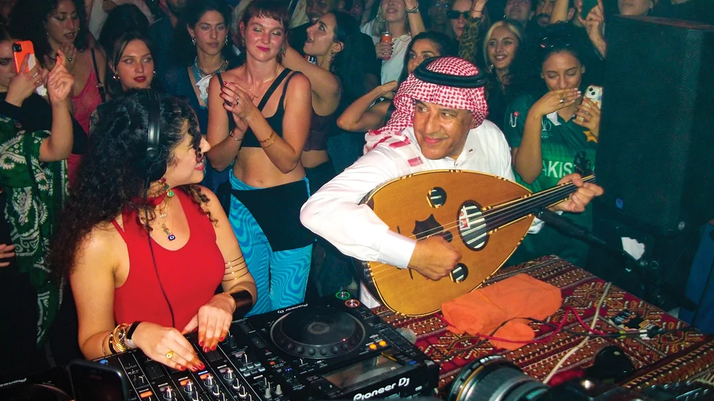 Photo from Nooriyah's Boiler Room set. She's smiling behind the decks wearing a red top. Her father is sat beside her playing the oud. He is wearing a Keffiyeh headdress 