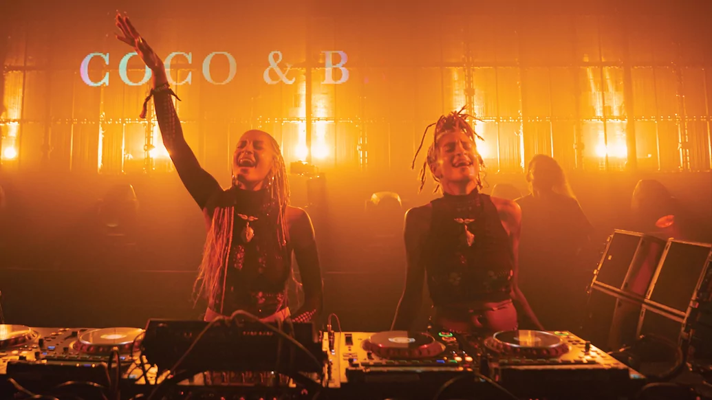 Photo of Coco & Breezy behind decks with orange lights in the background
