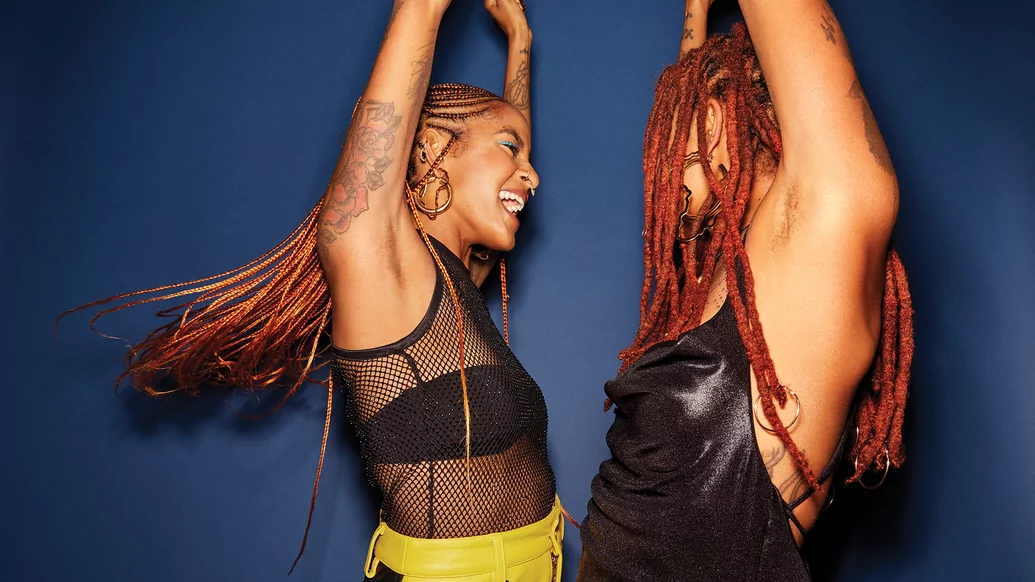 Photo of Coco & Breezy dancing with their hands in the air in front of a blue background