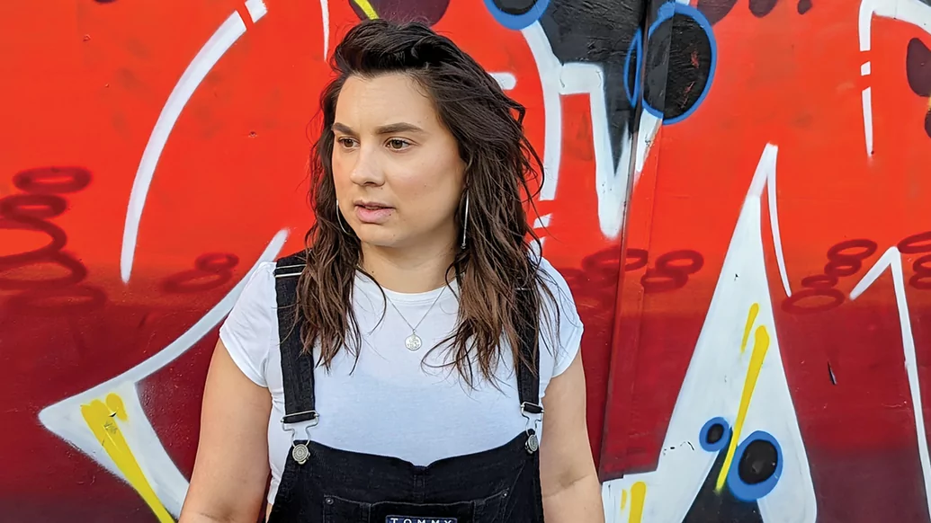 Photo of Amy B wearing a white t-shirt and black dungarees, standing in front of a graffiti background