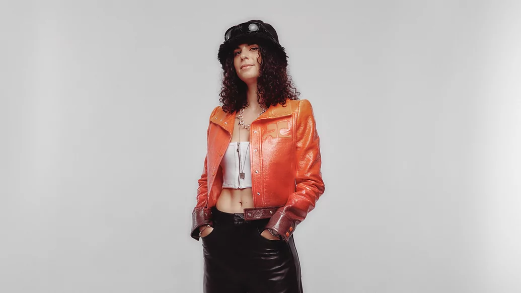 Photo of Carlita wearing an orange leather jacket and black skirt in front of a white background 