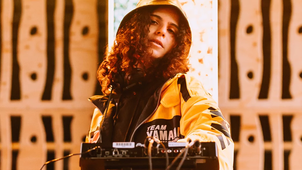 Photo of Carlita behind the decks while wearing a yellow and black jacket with a bucket hat