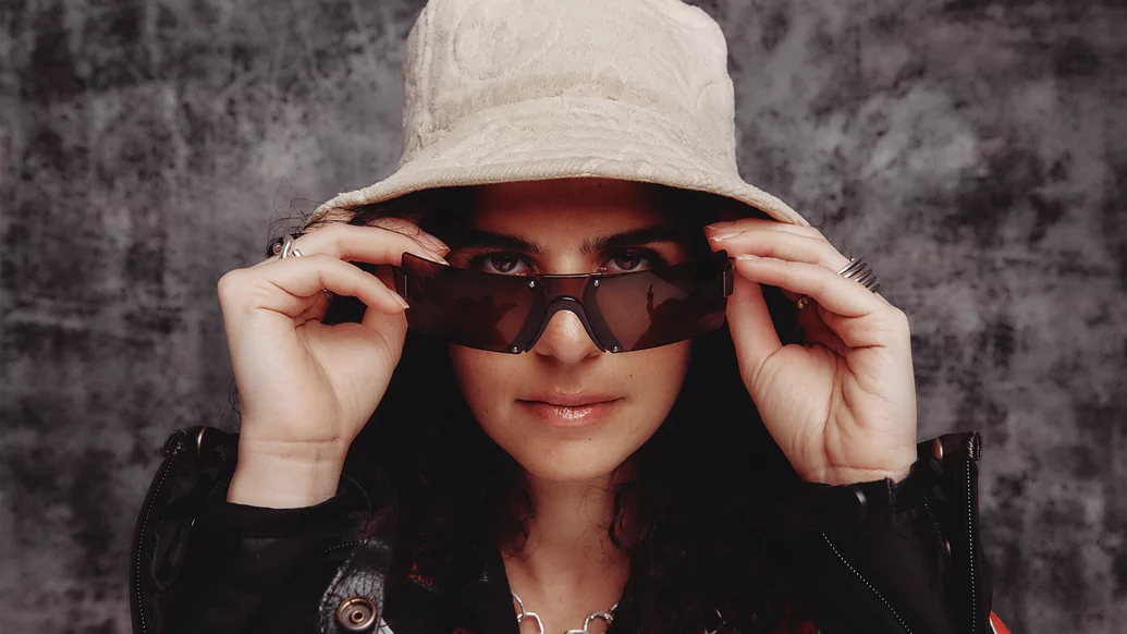Photo of Carlita posing wearing a pink bucket hat, black jacket, and black sunglasses which she is adjusting