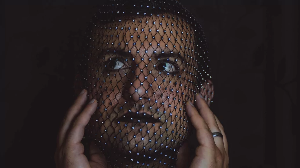Photo of Remniqe with a glittering mesh over their face against a black backdrop