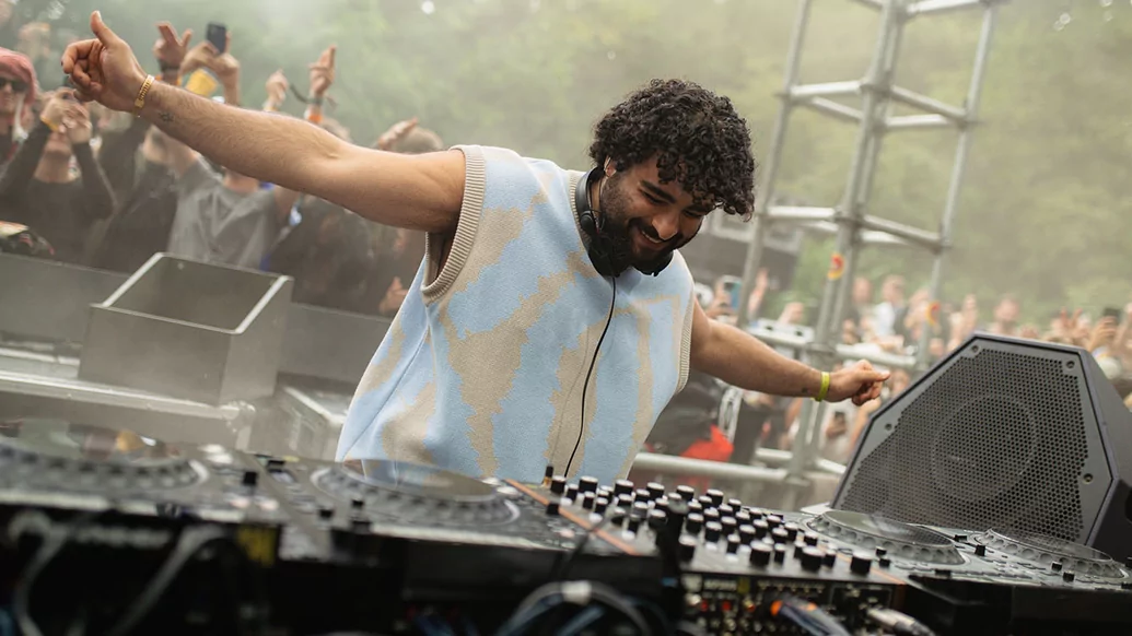 Fafi Abdel Nour DJing at Dekmantel festival wearing a blue and grey sleeveless wool vest. He's smiling with his arms outstretched
