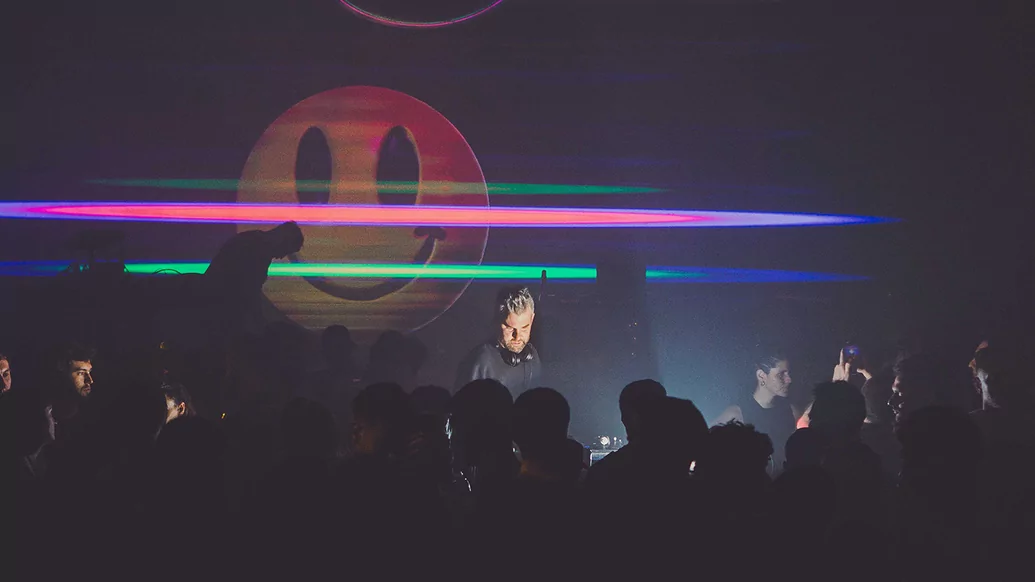 Voigtmann DJing in a busy, dark nightclub with a smiley face projected behind him