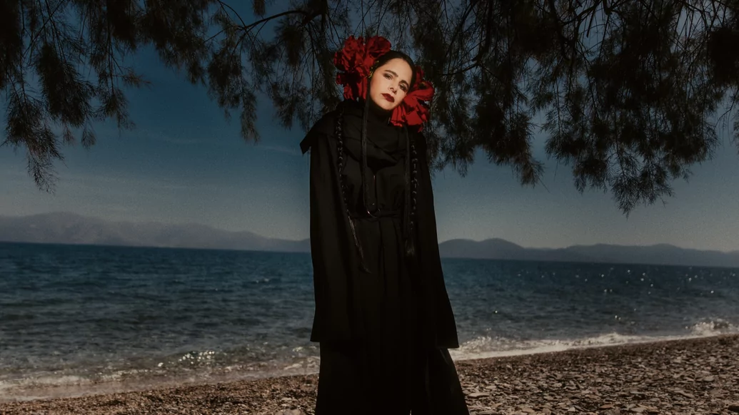 Photo of Sofia Kourtesis wearing a black cape with red neck detail on a beach
