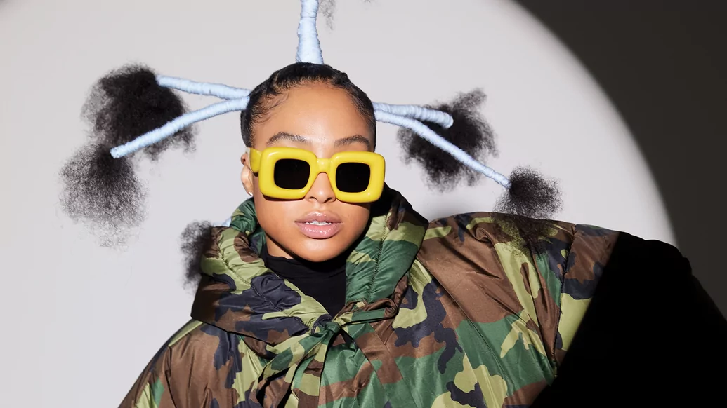 Photo of HoneyLuv wearing a camo puffer and yellow sunglasses with vertical braids in her hair 