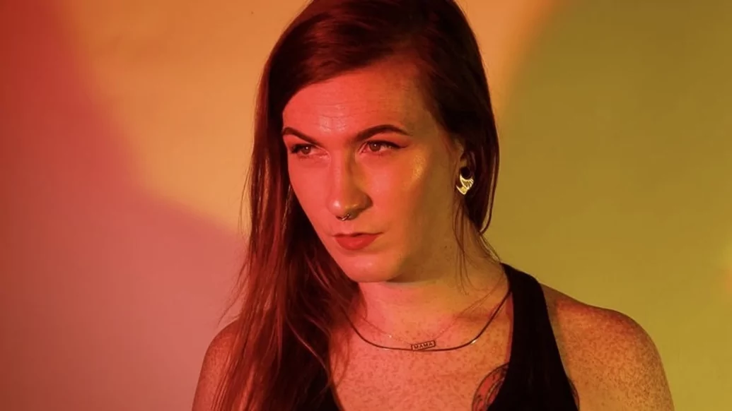 Photo of Mirin Doja wearing a black vest in front of a yellow and orange lit wall