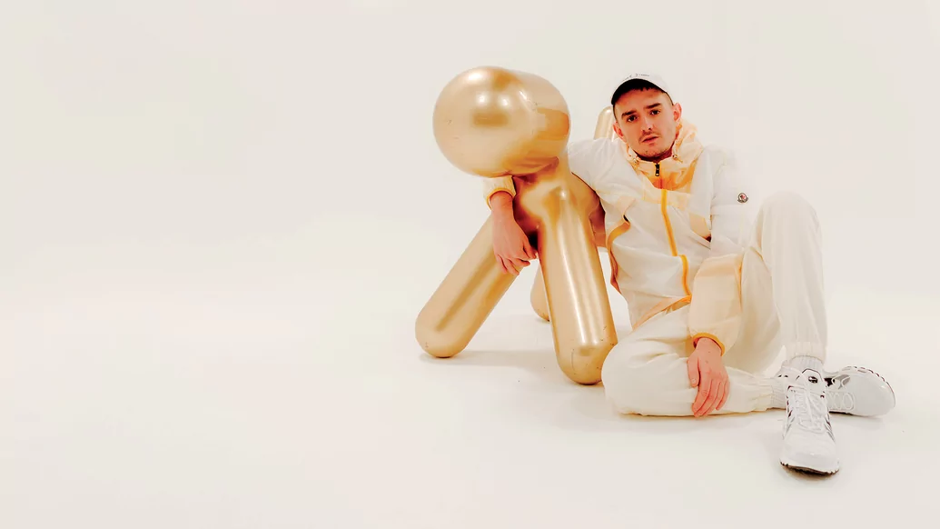 Photo of Ben Hemsley wearing a white tracksuit while sitting next to a golden dog statue