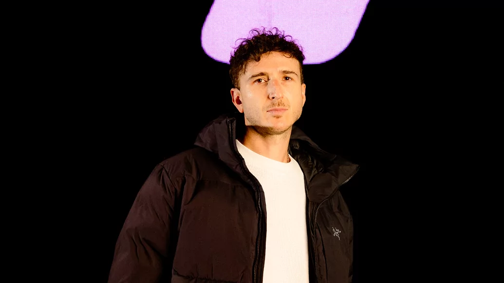 Photo of RL Grime wearing a black puffer and white shirt in front of a pink light