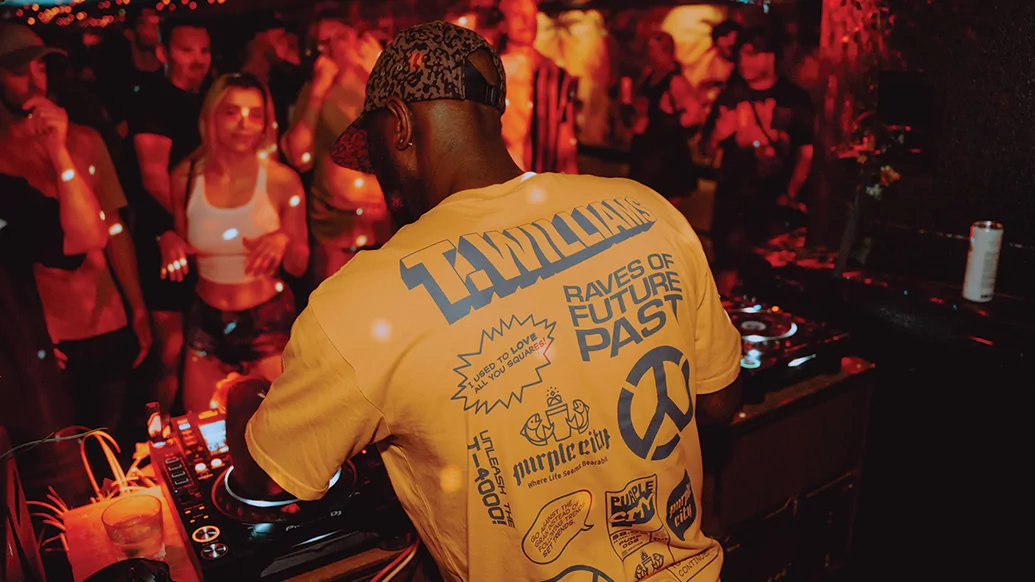 Photo of T.Williams DJing taken from behind. he's wearing a yellow t-shirt with the name of his debut album, 'Raves Of Future Past' on the back
