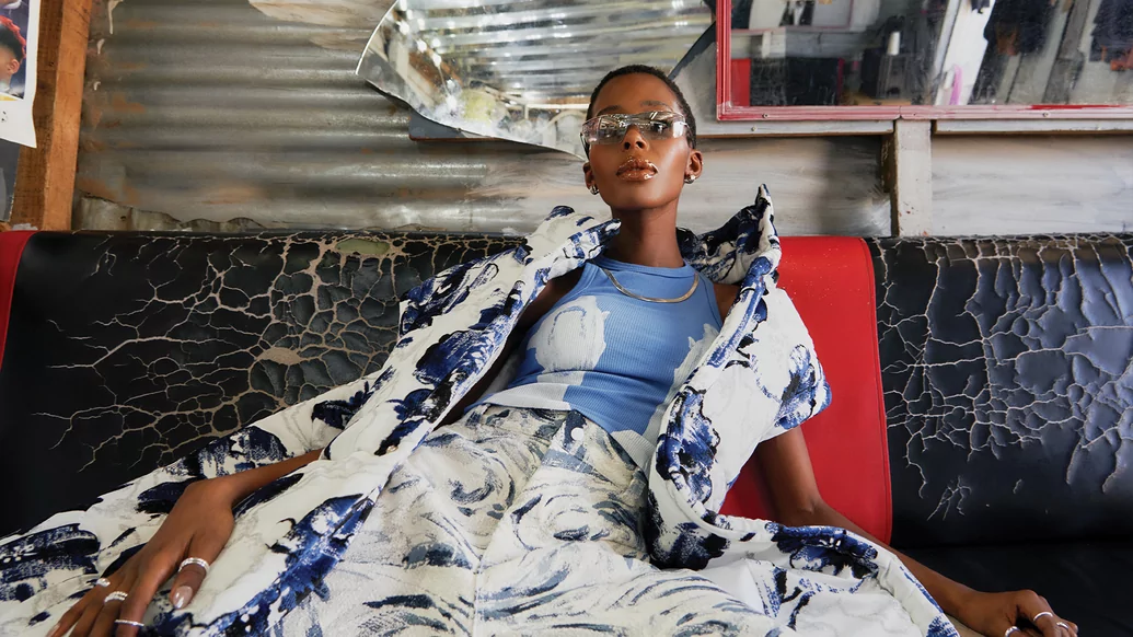Photo of DESIREE reclining on a black leather couch wearing a white trousers and jacket with an abstract blue pattern and shades