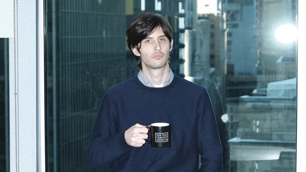 Photo of Plebeian wearing a blue jumper while holding a mug in front of an high-rise office background