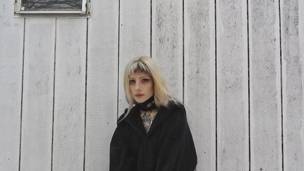 Photo of fknsyd standing against a white wooden wall wearing a big black coat