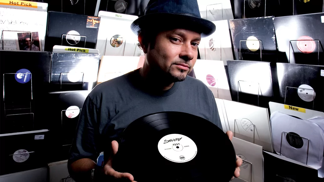 Photo of Louie Vega holding a vinyl surrounded by walls of records