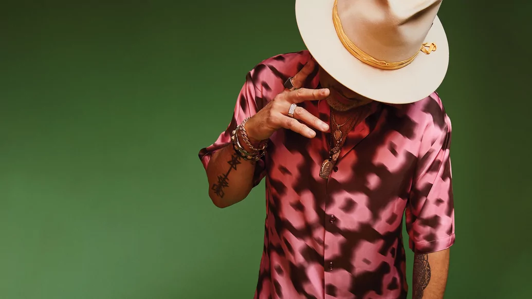 Photo of Louie Vega wearing a colourful flowery shirt and cream hat while posing against a green background
