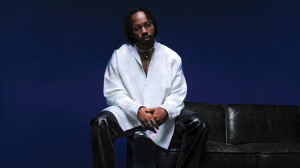 Photo of Channel Tres sitting on a black sofa whilst wearing a large white shirt