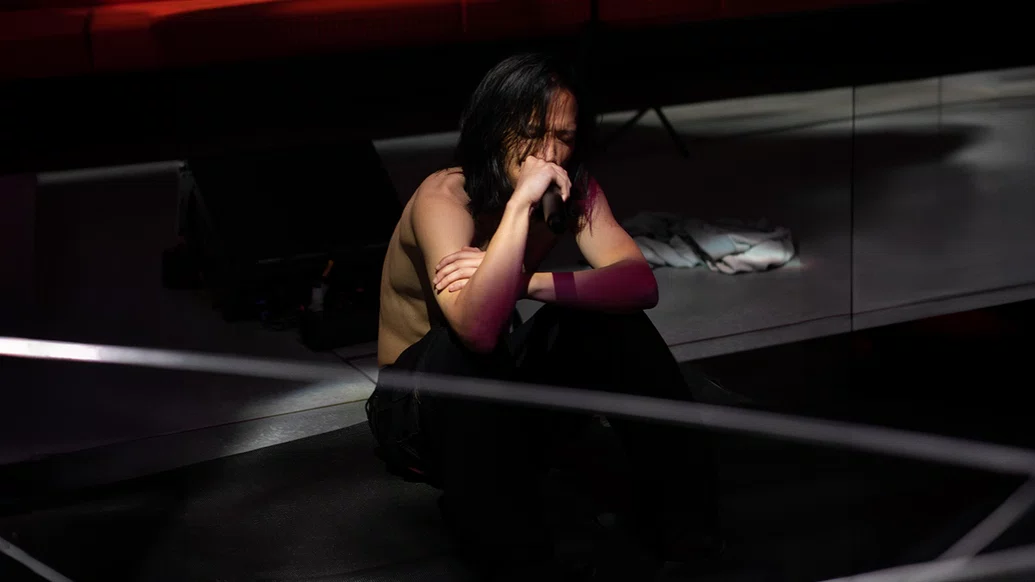 Lavurn sitting on a dark stage shirtless singing into a mic with his eyes closed