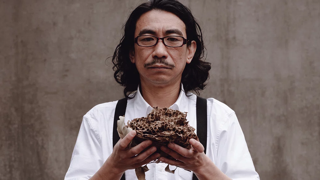 DJ Nobu wearing a white shirt, suspenders and glasses, holding a large shell in front of him with two hands