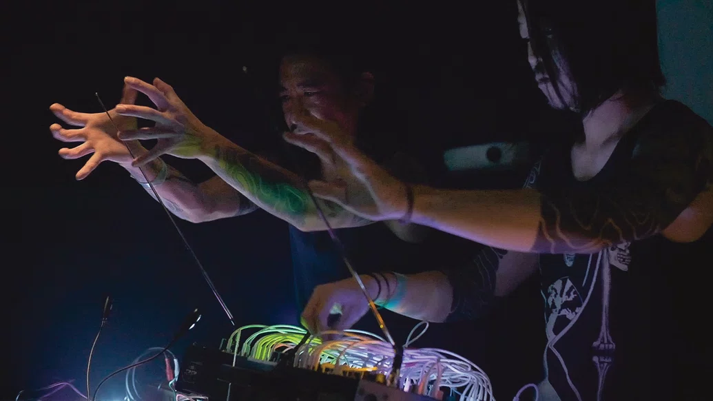 DJ Nobu and Doltz performing their Omega Point live set, their hands are held in clawlike formation over theremin arms