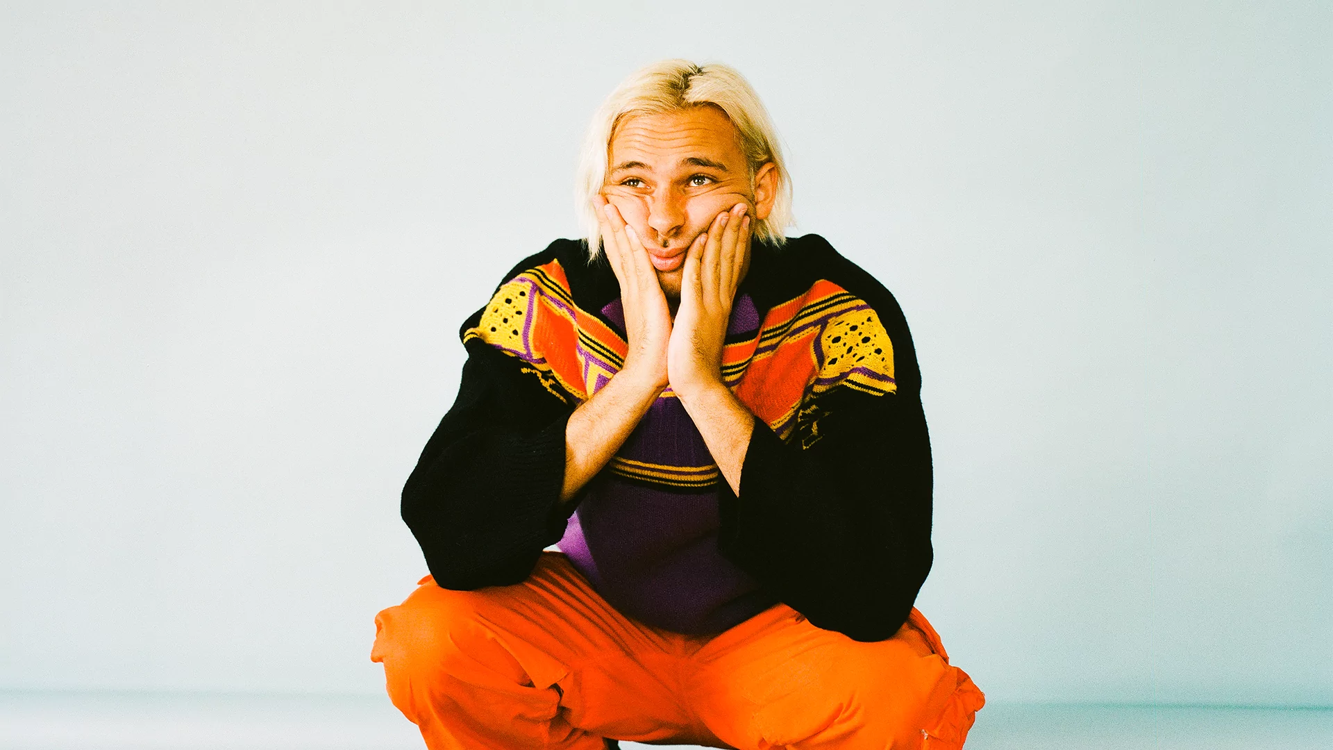 Flume resting his chin on his hands, in orange trousers and an orange and black jacket against a neutral background