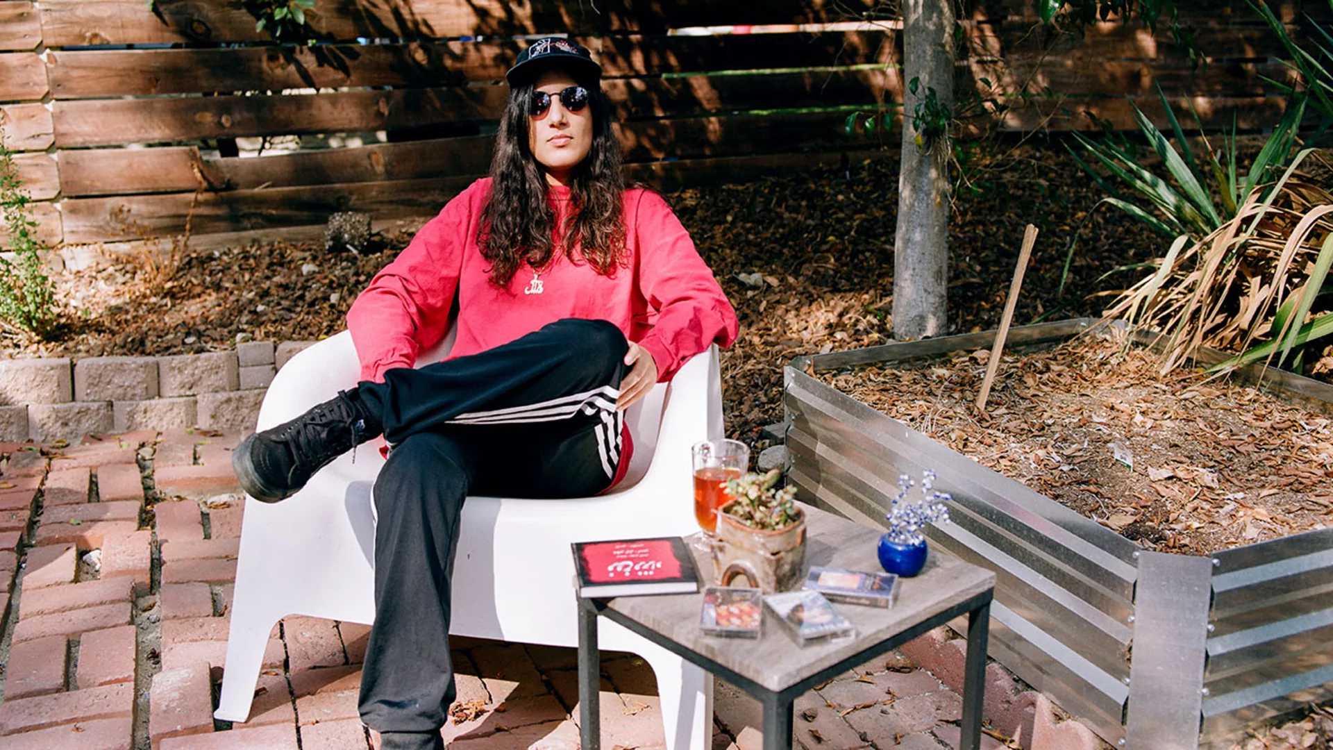 Maral in a red sweater, cap and sunglasses sitting on a white chair on a sun-lit patio