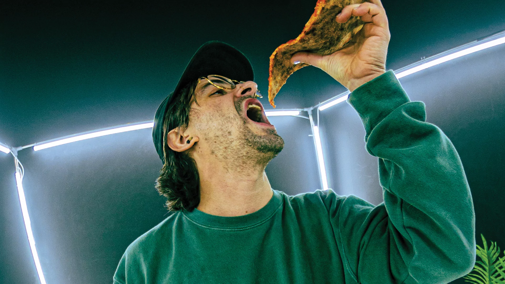 Devon James in a green sweater, cap and glasses, leaning back while eating a slice of pizza in front of white strip lights