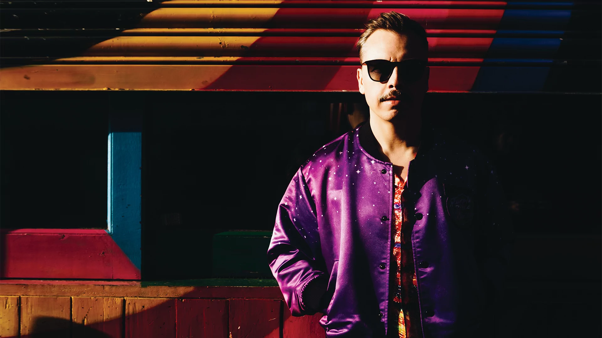 Purple Disco Machine wearing shades and a purple jacket in front of a multi-coloured 