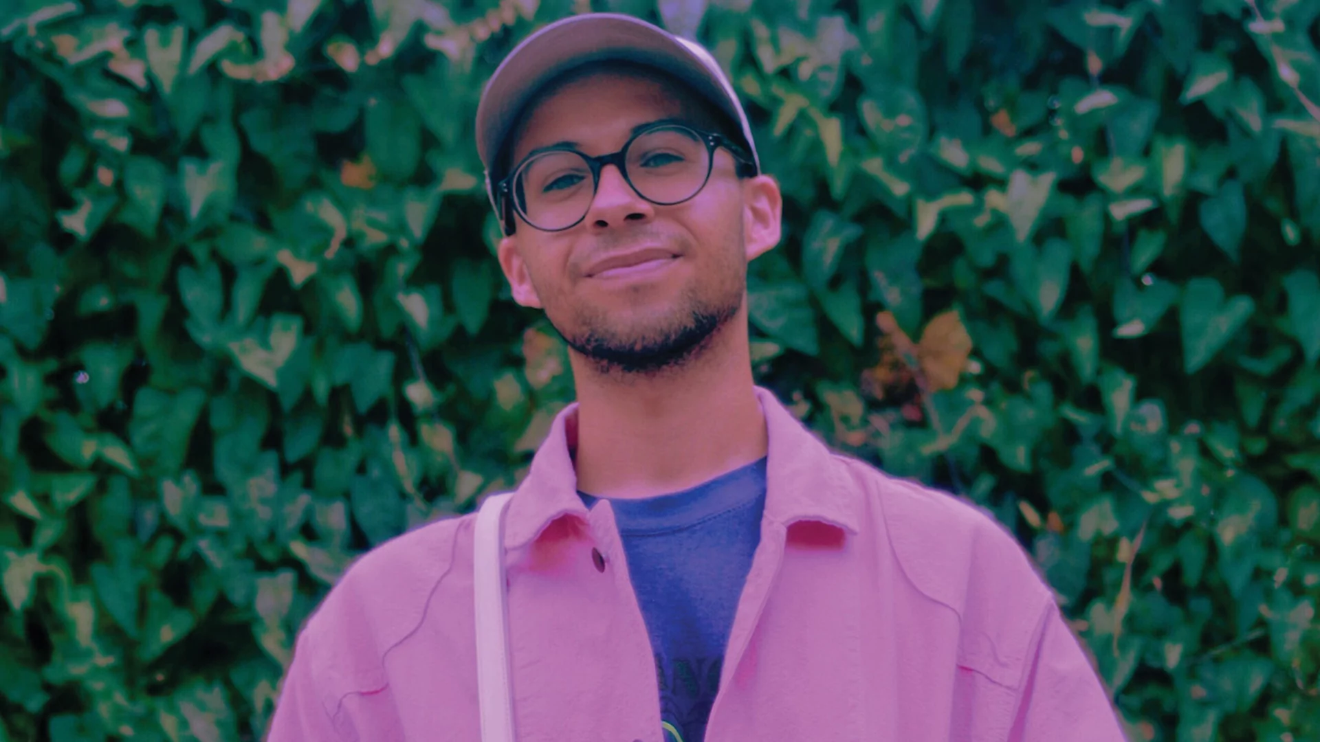 DJ SWISHA standing in front of a tall green hedge wearing a pink button up shirt, cap and glasses