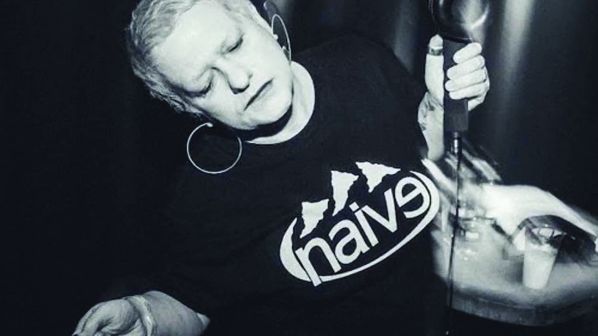 Black and white picture of Jaye Ward dancing while holding DJ headphones, wearing a Naive record label t-shirt
