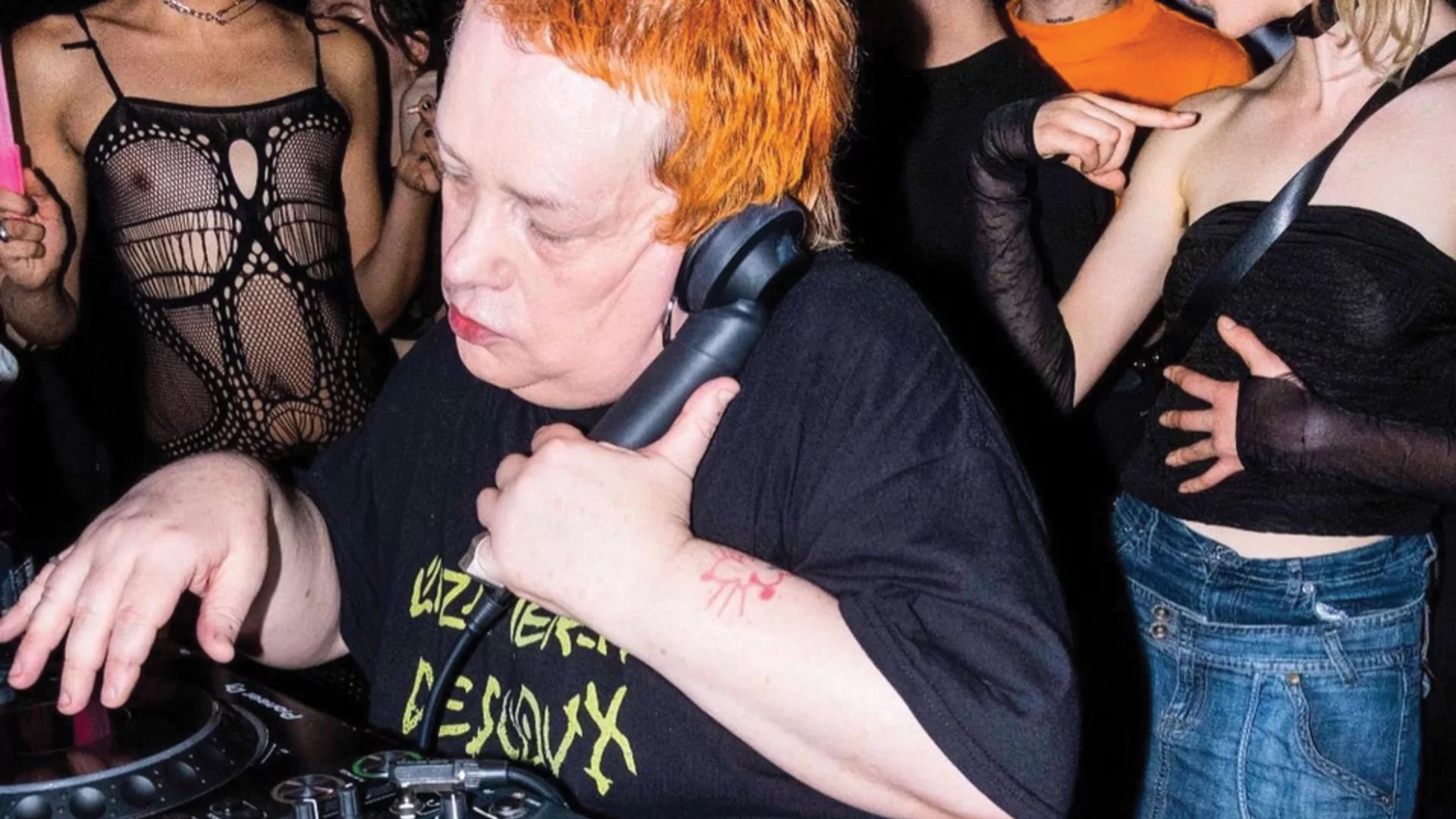Jaye Ward with short cropped orange hair DJing with a group of dancers behind her