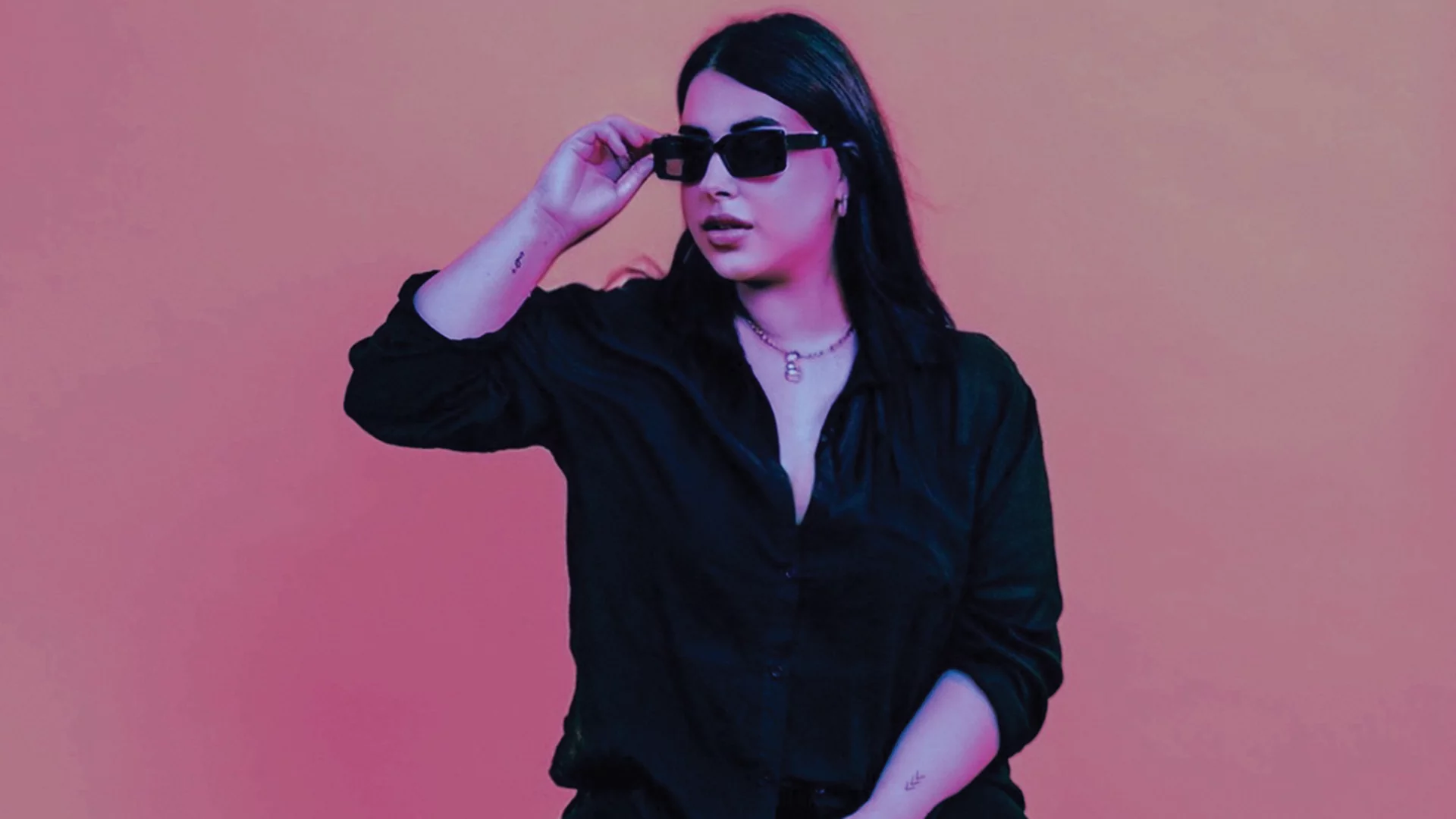 Naz wearing black sunglasses, standing against a pink backdrop