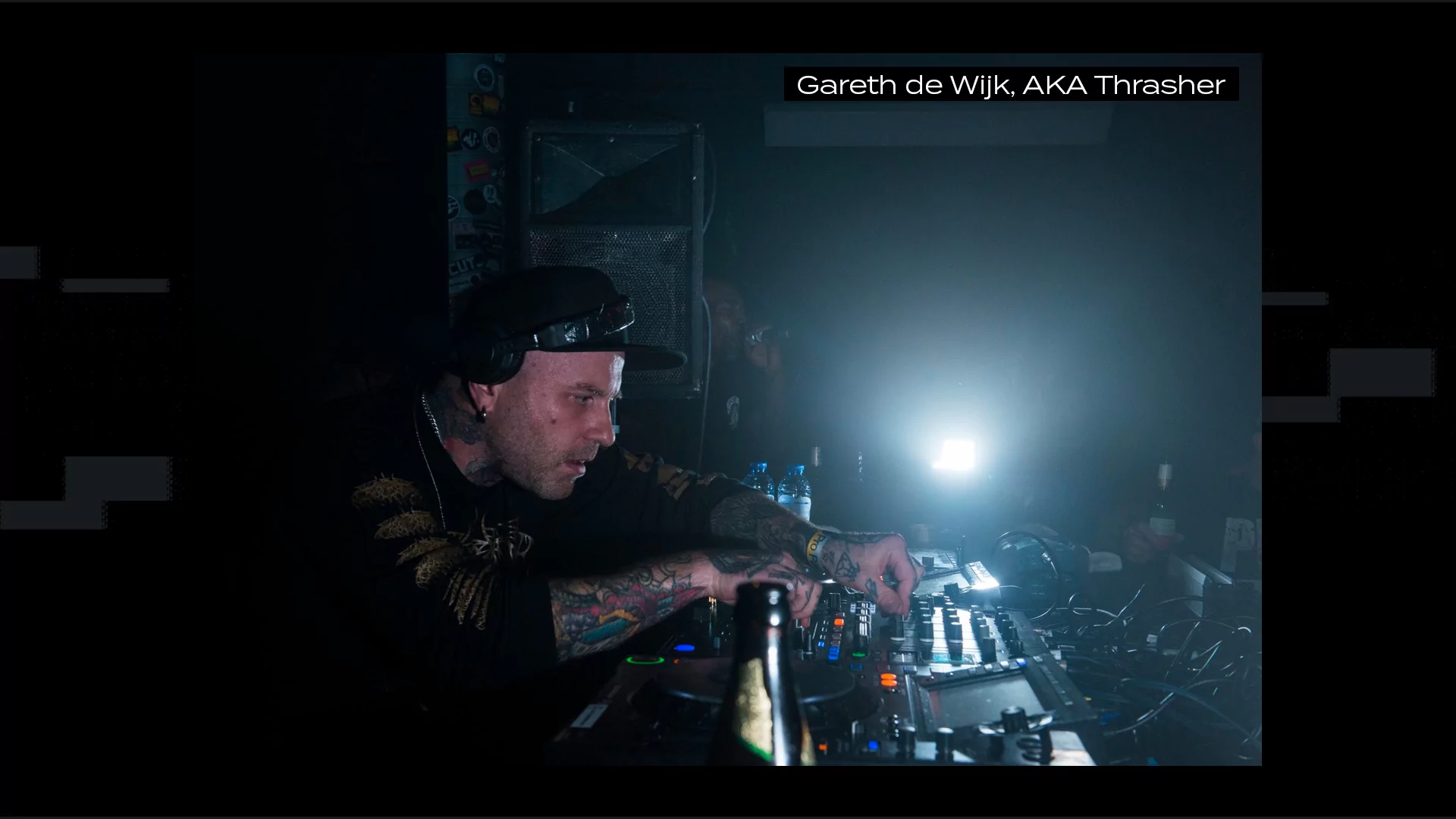 Photo of Gareth de Wijk, aka Thrasher, leaning over CDJs DJing. A single spotlight shines on him. He's wearing a baseball cap and is covered in tattoos