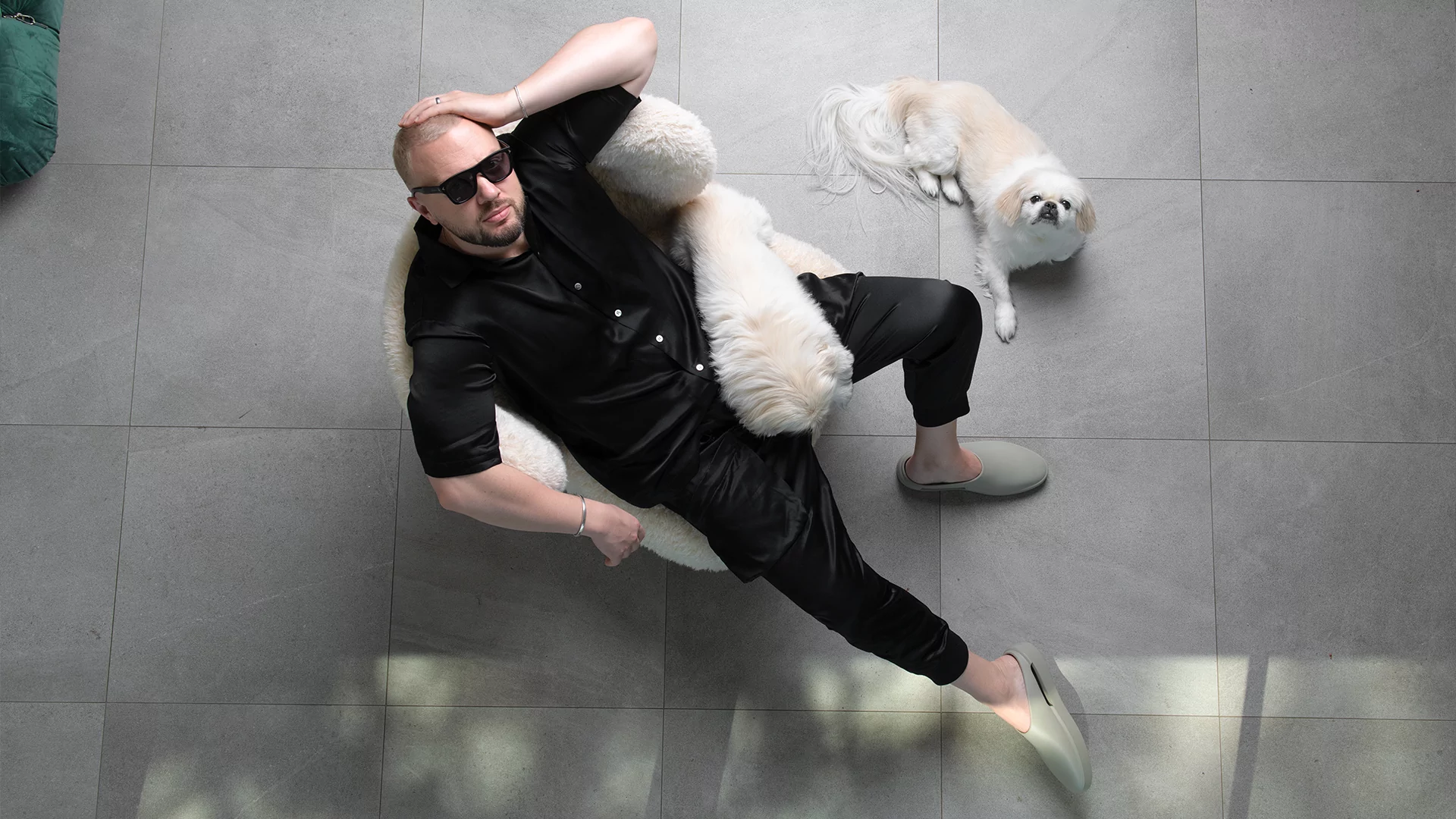 Photo of Chris Lake wearing all black and posing on a chair with two white dogs. 