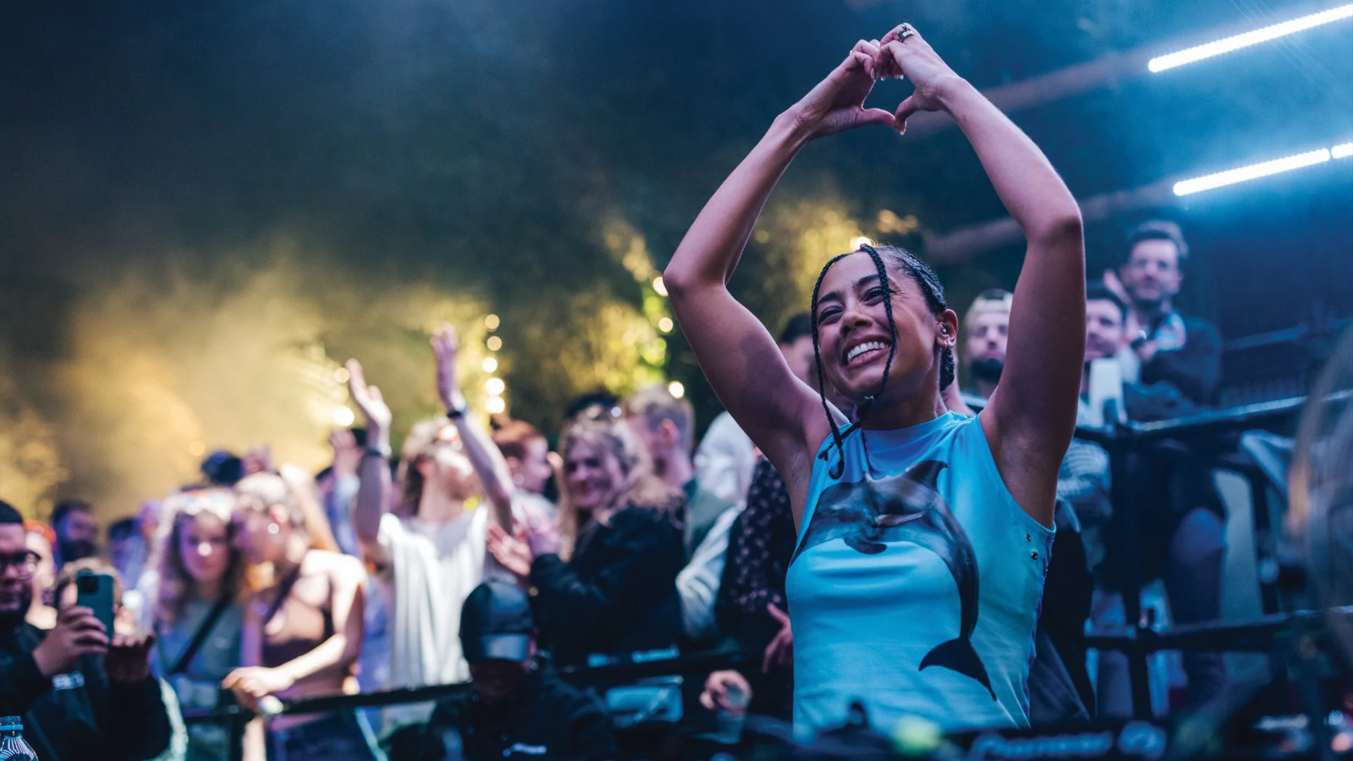 Photo of Jayda G DJing. She is grinning widely and has her hands above her head making a love heart shape. Some members of the crowd are dancing behind her