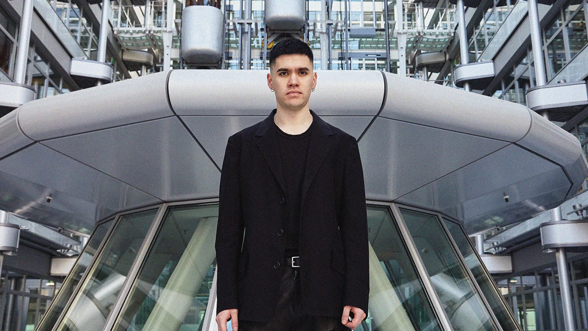 Photo of Phase Fatale standing in front of a tall modern building with many windows. He's wearing a black suit jacket and black t-shirt