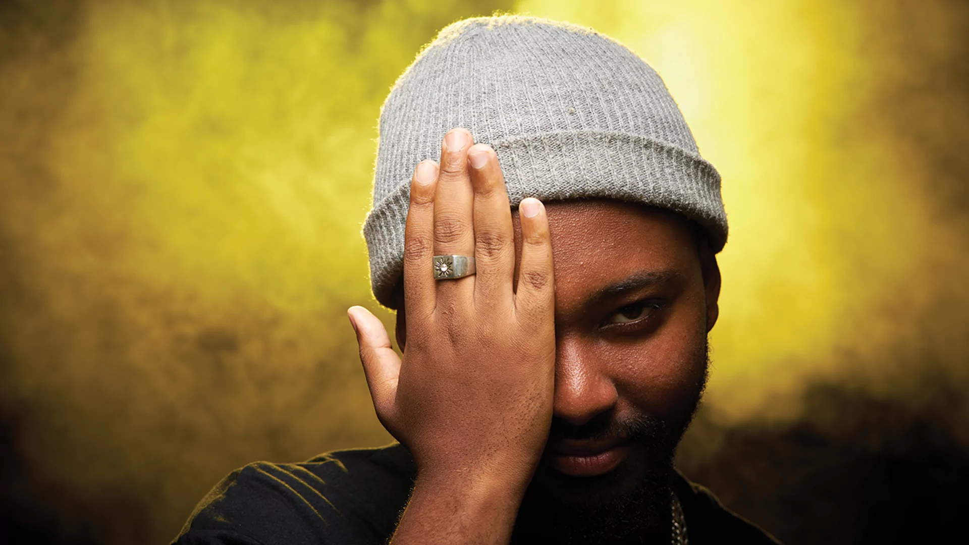 Thuto the Human's press shot. He's against a smoky yellow background and wearing a grey beanie hat. His right hand is covering his eye