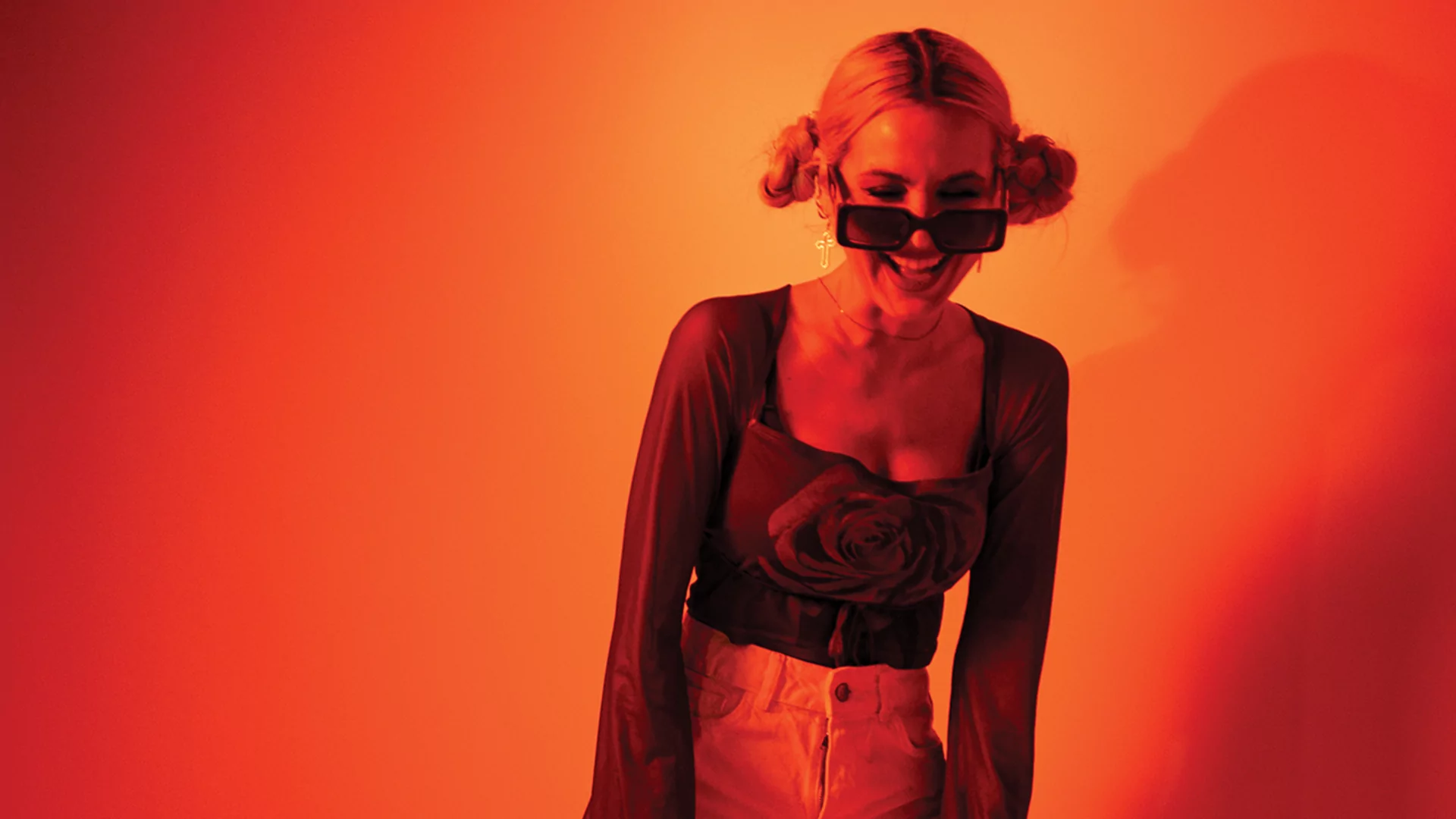 Leena Punks posing in a red and orange-lit room. She's laughing. Her hair is tied up in buns and she's wearing sunglasses
