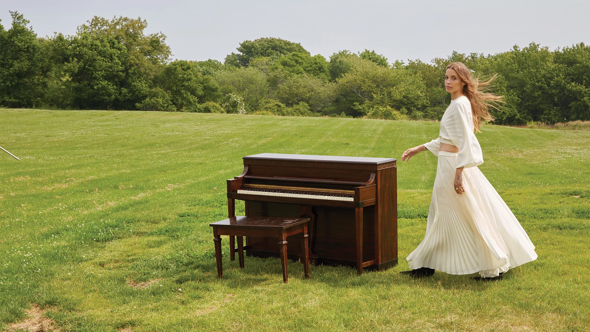 Photo of LP Giobbi posing next to a piano in a green field while wearing a flowing white dress