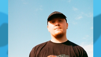 Chris Stussy in a black t-shirt and hat against a blue sky