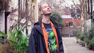 DJ Leisureware standing in an alley looking at the sky, wearing a raincoat and alien print tshirt
